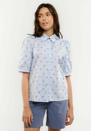 Naine, kes kannab helesinist lühikese varrukaga pluusi siniste südamemustritega ja siniseid lühikesi pükse, seisab ühetaustalise valge tausta ees.