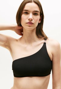 Sort bikini top med én skulder, lavet af tekstureret stof med en glat overflade og en dekorativ stropdetalje. Tætsiddende design uden polstring.