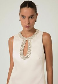 Bezramenné krémové šaty s výstrihom v tvare kľúča, zdobené korálkovým golierom. Hladká textúra s priliehavým strihom. Žiadne viditeľné vzory.