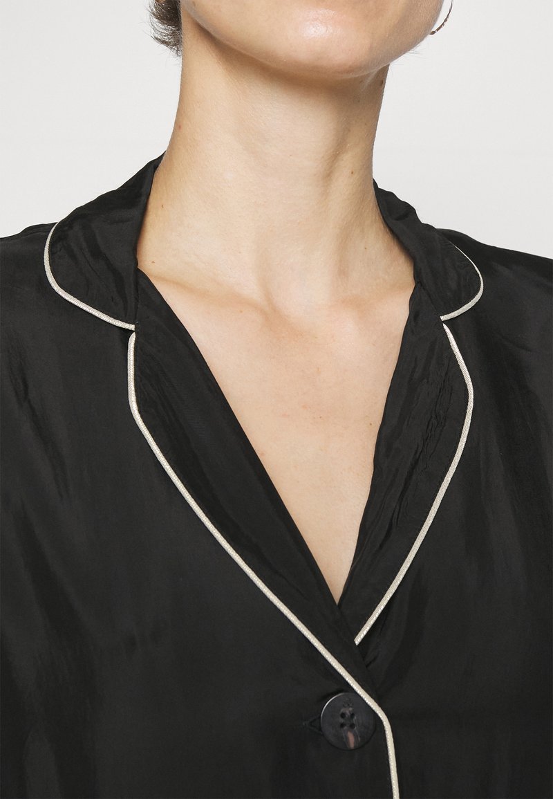 Zwarte zijde shirt met een sjaalkraag, contrasterende witte bies en één zwarte knoop. Gladde textuur, v-hals ontwerp.