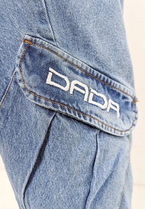 Helles blauer Denim-Stoff mit subtiler Textur; verfügt über eine abgerundete Taschenklappe mit weißem, gesticktem "DADA"-Logo und kontrastierendem orangefarbenem Stich.