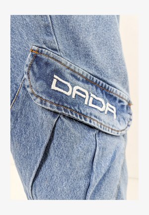 Helles blauer Denim-Stoff mit subtiler Textur; verfügt über eine abgerundete Taschenklappe mit weißem, gesticktem "DADA"-Logo und kontrastierendem orangefarbenem Stich.