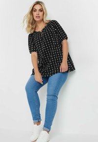Blusa nera con pattern geometrico bianco, vestibilità rilassata, maniche corte e scollatura con cordoncino, abbinata a jeans skinny blu e sneakers bianche.