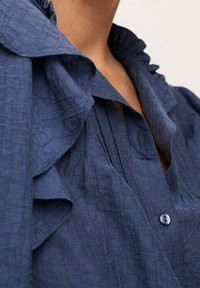 Navy blue blouse with a textured fabric, featuring a ruffled collar and placket, and buttons down the front.