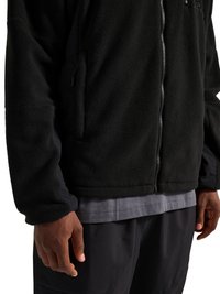 Sort fleecejakke med lynlås, båret over en grå skjorte og sorte bukser, vist fra midt på torsoen til hænderne på en person.