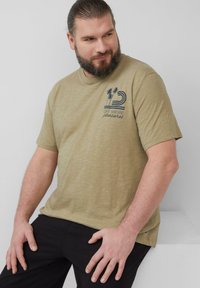 Olijfgroene T-shirt gemaakt van lichte stof met een ronde halslijn, voorzien van een blauwe grafische print van palmbomen en golven op de borst.