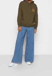 Felpa verde oliva con tasca frontale e dettagli con logo; abbinata a jeans in denim blu a gamba larga e sneakers bianche.