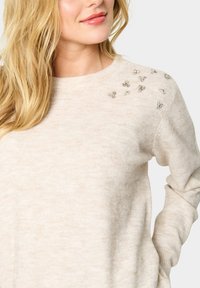Šviesiai rudas megztinis su apvalia apykakle, minkšta tekstūra ir dekoratyviniais kristalų akcentais kairiajame petyje, pabrėžiantis paprastą ir elegantišką dizainą.