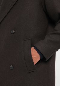 Mano inserita nella tasca di un cappotto di lana marrone scuro con due bottoni neri visibili, mostrando la manica del cappotto e il polsino blu navy sottostante.