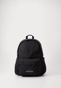 Sac à dos en nylon noir de forme arrondie, avec une poche zippée à l'avant et une poignée supérieure. Présente le logo Calvin Klein en blanc sur le devant.