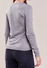 Kobieta zwrócona tyłem, ubrana w jasnoszary sweter z długim rękawem i ciemnoniebieskie dżinsy, na jednolitym beżowym tle.