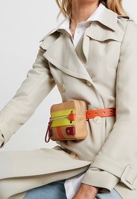 Kvinna som bär en beige trenchcoat med ett orange bälte och en liten bruna, röda och gula dragkedjepåse fäst vid midjan.