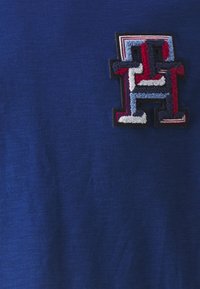 Modré bavlněné tričko s vícebarevným vyšívaným logem na levé straně hrudi, s texturami v červených, bílých a modrých vláknech.