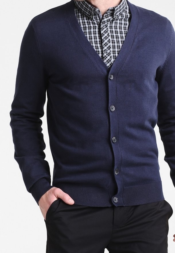 Homme portant un cardigan bleu marine boutonné par-dessus une chemise à carreaux noir et blanc, une main dans la poche d'un pantalon noir.