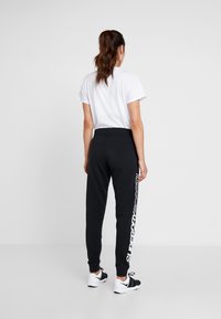 Schwarze Jogginghose mit elastischem Bund und gerippten Bündchen, mit einem weißen vertikalen Logoaufdruck auf dem linken Bein. Kombiniert mit einem weißen T-Shirt.