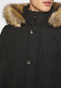 Schwarze Parkajacke mit einem fellbesetzten braunen Kapuzenrand, ausgestattet mit einem Reißverschluss und Druckknöpfen sowie einer glatten Stoffstruktur. Zwei sichtbare Fronttaschen.