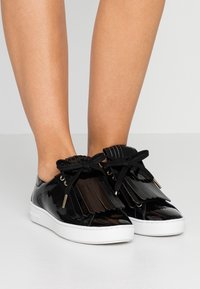 Glänzende schwarze Sneaker mit weißen Sohlen, schwarzen Schnürsenkeln und dekorativem schwarzen Fransen oben, barfuß vor einem schlichten Hintergrund getragen.