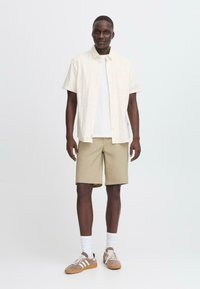 Lys beige kortærmet skjorte, hvid undertrøje, khaki shorts og grå sneakers med hvide accenter og snørebånd. Enkel design, afslappet pasform.