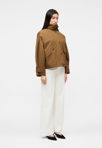 Γυναίκα που στέκεται φορώντας καφέ παλτό με ψηλό γιακά, λευκό παντελόνι φαρδύ στο κάτω μέρος και μαύρα μυτερά παπούτσια, μπροστά σε απλό φωτεινό φόντο.