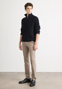 Pull en maille côtelée marine avec col montant, associé à un pantalon beige et des chaussures en cuir noires, se tenant sur un sol aux tons neutres.