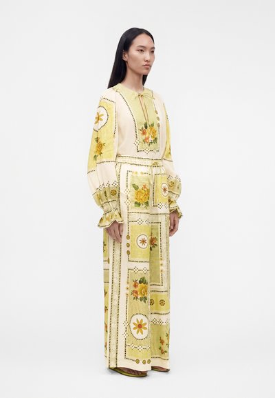 Γυναίκα που στέκεται πλαγίως, φοράει ένα μακρύ κρεμ φόρεμα με κίτρινα λουλουδάτα και γεωμετρικά σχέδια, φουσκωτά μανίκια και πράσινα σανδάλια.