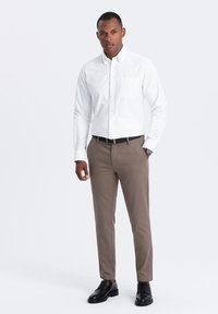 Witte lange mouwen blouse met knopen en zak, gecombineerd met lichtbruine slim-fit broeken. Zwarte leren schoenen. Neutrale achtergrond.