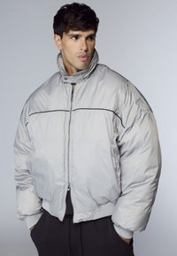 Giovane uomo che indossa un giubbotto imbottito grigio chiaro con colletto alto e pantaloni neri, che guarda a sinistra con le mani in tasca.