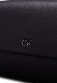 Černý texturovaný kožený povrch se stříbrným logem "CK" uprostřed módního doplňku, pravděpodobně kabelky nebo peněženky.