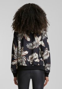 Veste bomber noire avec un motif de feuilles tropicales en crème et marron, avec un ourlet et des poignets côtelés, et un tissu à la texture lisse.