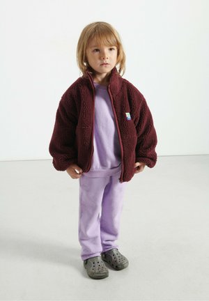 Petit enfant portant une veste en polaire bordeaux, un sweat-shirt violet clair et un pantalon, ainsi que des chaussures à enfiler gris foncé, se tenant sur un sol clair.