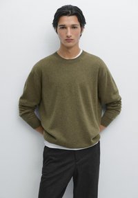 Maglione lavorato a maglia verde oliva con collo rotondo, vestibilità rilassata e polsini a costine, indossato sopra una camicia bianca, abbinato a pantaloni grigio scuro.