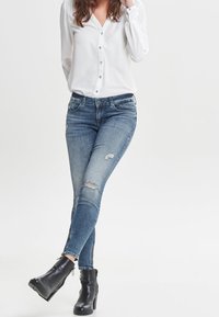Jeans in denim azzurro chiaro con dettagli sfrangiati e leggera scoloritura, abbinati a una camicia bianca con bottoni e stivaletti neri alla caviglia.