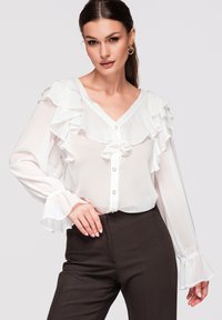 Femme portant un chemisier blanc à volants avec des boutons dorés et un pantalon taille haute foncé, posant devant un fond clair uni.
