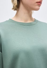 Νεαρό άτομο με κοντά ξανθά μαλλιά φοράει μια ανοιχτοπράσινη μπλούζα με στρογγυλή λαιμόκοψη μπροστά σε μια απλή φόντο.