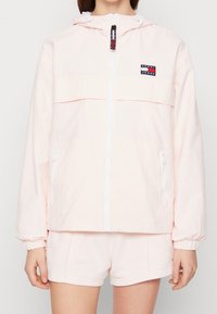 Jasnoróżowa kurtka wiatrówka z przednim zamkiem błyskawicznym, białymi akcentami, kapturem i naszywką z logo na piersi. Posiada elastyczne mankiety i boczne kieszenie.