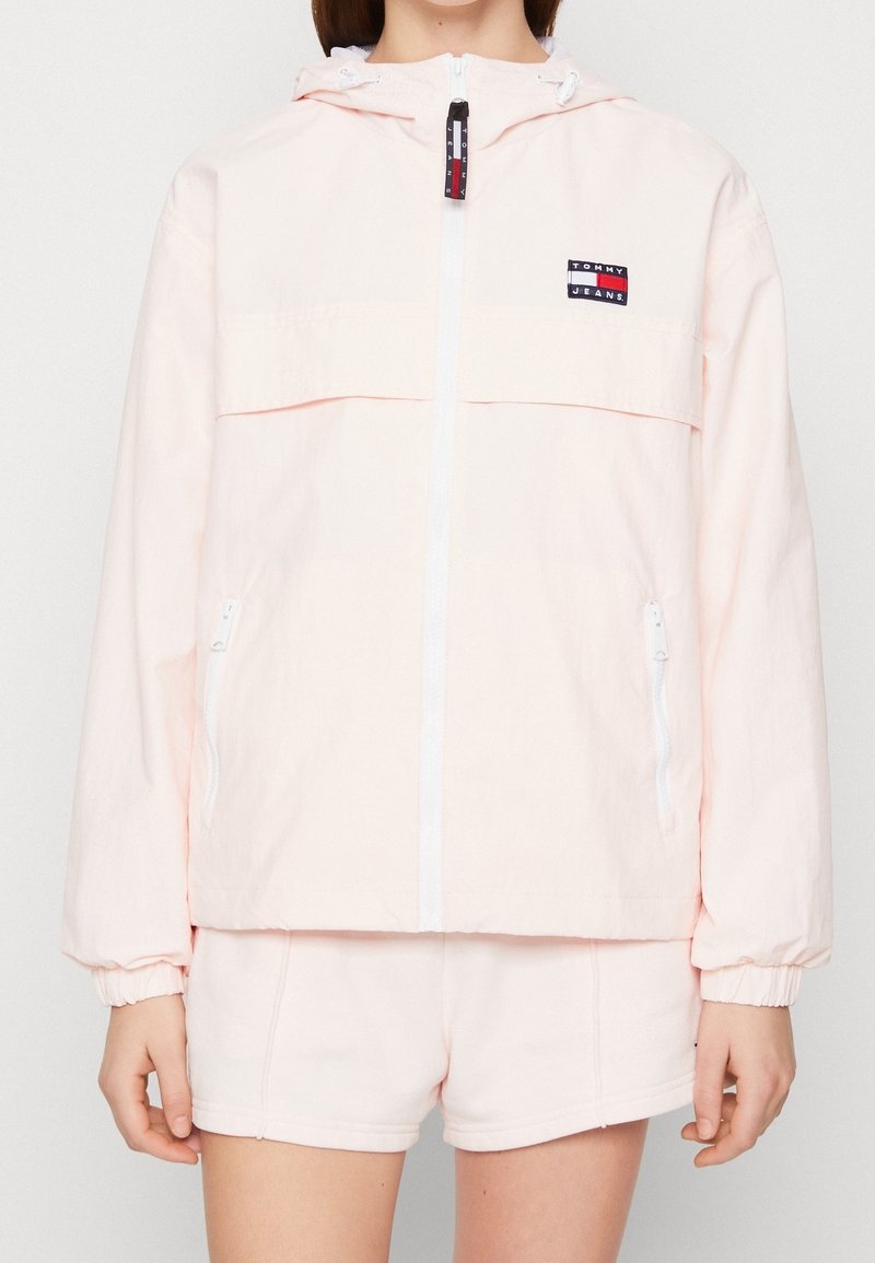 Jasnoróżowa kurtka wiatrówka z przednim zamkiem błyskawicznym, białymi akcentami, kapturem i naszywką z logo na piersi. Posiada elastyczne mankiety i boczne kieszenie.