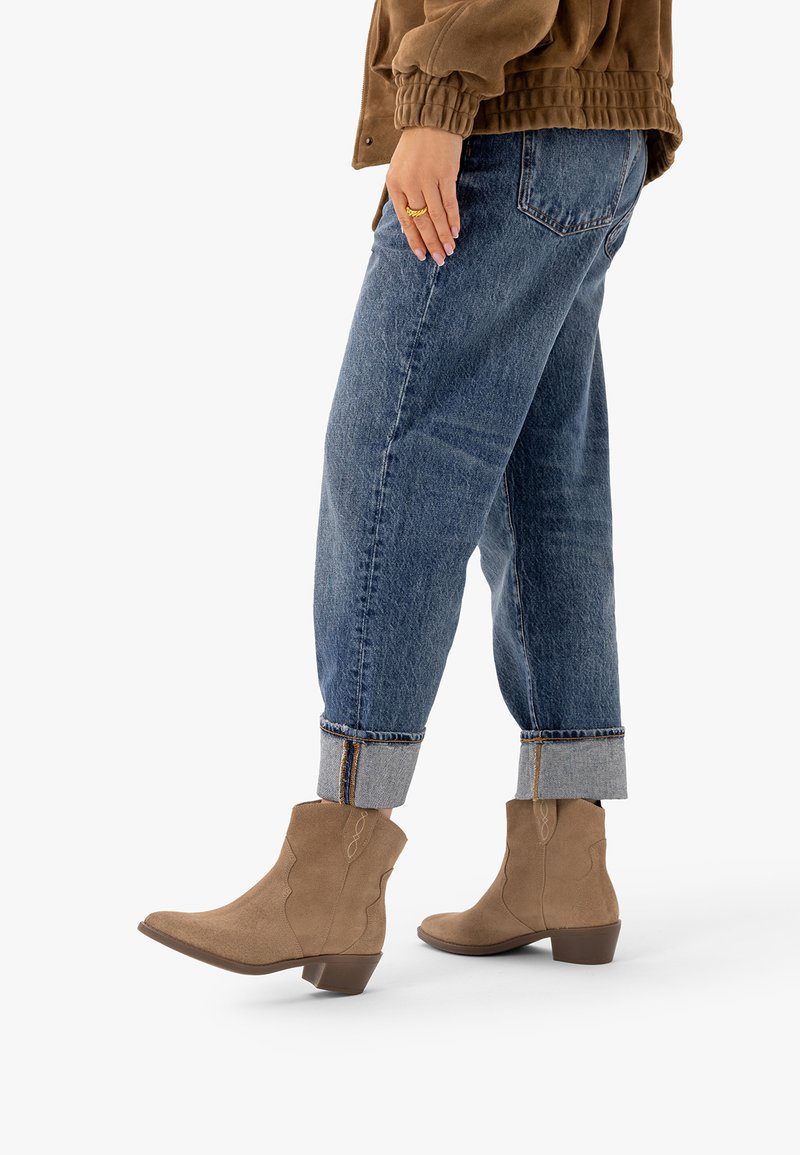 Personne portant un jean bleu retroussé, des bottines marron en daim à talons bas et une veste marron, debout de profil sur fond blanc.