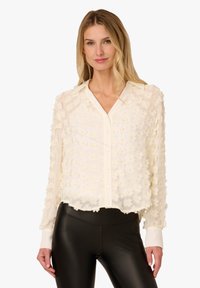 Woman wearing a sheer white button-up blouse with floral appliqués and black faux leather leggings, standing against a white background.