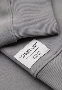 Poignet en tissu gris côtelé avec une étiquette rectangulaire blanche portant les inscriptions « YOUR ONLY LIMIT IS YOUR MIND » et « OMBRE depuis 1985 » cousue dessus.