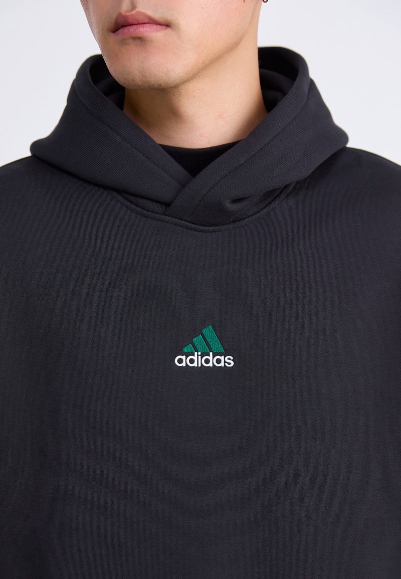 Primer plano de una persona que lleva una sudadera con capucha negra con un logo de Adidas en verde y blanco en el pecho, contra un fondo liso.