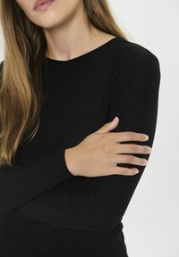 Woman with long brown hair wearing a black long-sleeve top, crossing her arm and holding her opposite upper arm against a plain background.