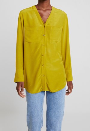 Personne portant une chemise ample jaune moutarde à boutons avec poches poitrine et un jean bleu clair sur un fond uni.