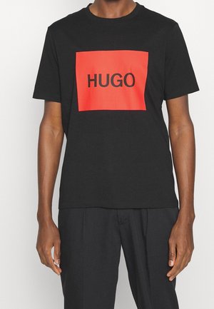 Schwarzes Baumwoll-T-Shirt mit einem großen roten rechteckigen Logo, auf dem "HUGO" in fetten schwarzen Buchstaben steht, kurzen Ärmeln und einem Rundhalsausschnitt.