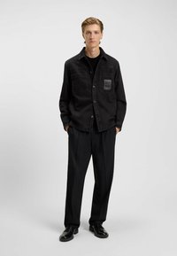Zwarte denimshirt met twee voorzakken en een leren patch, gecombineerd met zwarte op maat gemaakte broek en leren schoenen, wat een monochrome look creëert.