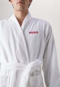 Weißer Frottee-Bademantel mit Schalkragen, vorderen Taschen und gebundenem Gürtel; auf der linken Seite befindet sich ein rotes, gesticktes "HUGO"-Logo.