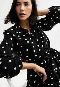 Schwarze Bluse mit Punkten, Knopfverschluss an der Vorderseite, elastischen Puffärmeln und einem gerafften Taillendetail. Glattes Material mit einer weichen Textur.