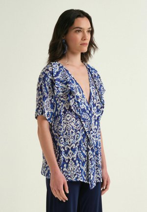 Woman wearing a blue and white patterned blouse with ruffled V-neck, short sleeves, and dark blue pants, standing against a plain light background.