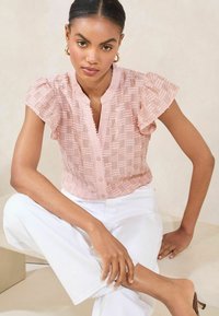 Blusa rosa testurizzata con maniche corte a volant, chiusura frontale con bottoni e motivo sottile abbinata a pantaloni bianchi. Tessuto morbido, vestibilità casual.