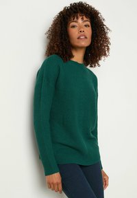 Grüner Strickpullover mit Rundhalsausschnitt, gerippter Textur und überschnittenen Schultern, kombiniert mit dunklen Jeans.