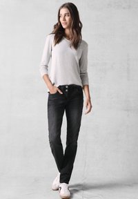 Grijze longsleeve top, zwarte jeans met twee-knopensluiting, skinny fit, witte sneakers, zelfverzekerd staand voor een effen achtergrond.
